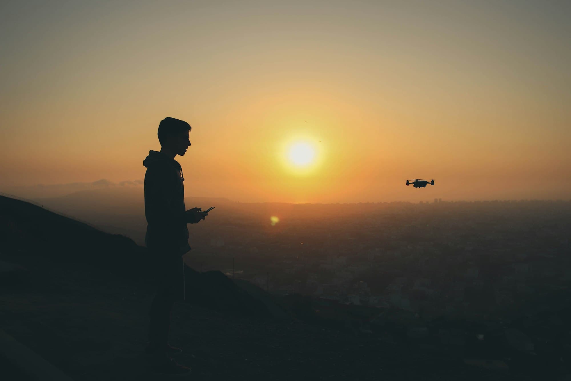 A drone pilot silhouetted against the setting sun, flying above a valley